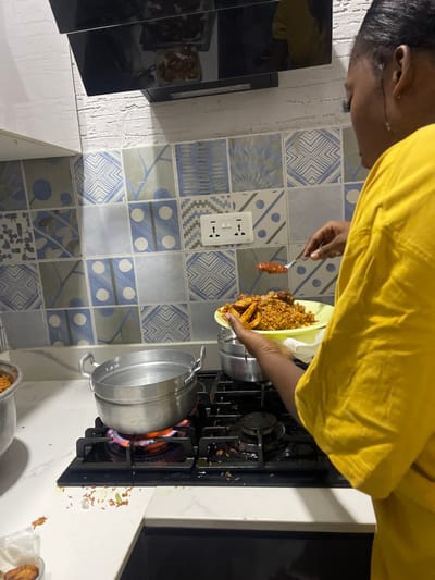 The image depicts a person in a kitchen, engaged in the act of cooking or serving a meal. The individual, visible from the chest up and wearing a yellow t-shirt, is holding a light green plate laden with Jollof rice, fried plantains (dodo), and possibly chicken pieces. Their other hand holds a fork with an orange-colored piece of food, potentially another piece of plantain or a sauce component, positioned over the plate.
The kitchen setting features a gas stove with four burners, where a metal pot is actively heating over a blue and orange flame on the front-right burner. A second metal pot is partially visible in the bottom right corner. The wall behind the stove is adorned with a variety of blue, white, and grey patterned tiles, showcasing geometric, abstract, and subtle floral designs. A white electrical outlet with two sockets is mounted on the tiled wall. A clean white countertop is visible beneath the stove and to the left. Scattered around the gas burners are visible spills or debris, likely rice grains and other food particles, suggesting recent cooking activity.
This scene provides a glimpse into a domestic kitchen, likely within Adenta Municipality, Ghana, where a popular local dish featuring Jollof rice and plantains is being prepared or plated. The lighting indicates an indoor setting. No specific text is clearly decipherable within the image.