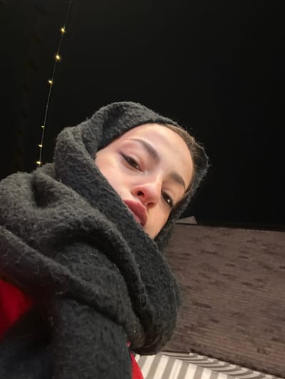 A woman wrapped in a charcoal-colored, textured scarf or blanket looks up at the camera from a low angle. Her face is partially obscured by the scarf, revealing her eyes, nose, and lips. The background is dark, with a brick building visible on the right and a string of small, warm-toned lights blurred in the lower left. The lighting suggests an outdoor nighttime setting.