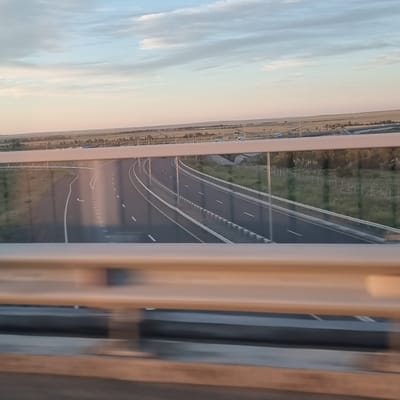 The image, rotated to present a horizontal view, depicts a highway with multiple lanes curving through a landscape. The road surface is dark asphalt, marked with white lines. A metallic barrier runs alongside the highway. To the left of the road, a grassy field extends towards a distant horizon. The sky above is a soft blend of blue and pale yellow, with wispy clouds, suggesting either early morning or late afternoon light.
On the far left of the image, a more distant view shows a treeline and some structures that appear to be houses or buildings, indicating a rural or semi-rural setting. The foreground is partially obscured by what seems to be the interior of a vehicle or building, with vertical elements suggesting framing or furniture.
The overall impression is one of travel, with the highway stretching into the distance under a serene sky. The time of day is likely either sunrise or sunset due to the lighting and sky colors. The location context provided (Alğa, Kazakhstan) suggests this is a highway in that region. No people or specific events are clearly visible. The image is devoid of any visible text.