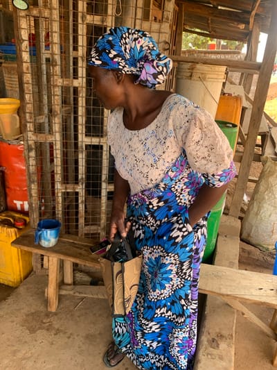 An adult female is depicted standing in a semi-outdoor commercial setting in Ube, Nigeria. She wears a blue, black, and white patterned headwrap and a matching long skirt or dress, combined with a white-cream lace short-sleeved top. Her right hand holds a small, light brown woven bag with black handles. She wears dark flip-flops. Her gaze is directed downwards and slightly to her left. The immediate background features a rusted metal grate structure. To her left, a yellow plastic funnel rests atop a partially visible red metal barrel marked "TI". A small, light brown wooden bench with a blue and white speckled mug is situated at floor level to her left. To her right, a large white plastic bucket, an orange plastic jerrycan, and a green plastic jerrycan are visible. The scene is structured by rudimentary wooden posts and beams, and the ground is an uneven, dirt-colored surface.