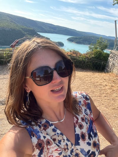 A woman is taking a selfie in a scenic outdoor location, likely a viewpoint overlooking a body of water. The woman has brown hair and is wearing large black sunglasses, a floral print dress with a V-neck, a silver necklace, and dangle earrings. Her mouth is slightly open as if speaking or smiling. In the background, a large turquoise lake is visible, surrounded by lush green hills. The sky is blue with some scattered clouds. The foreground consists of dry, light-colored ground and a stone wall on the right side of the frame. A metal railing runs along the edge of the viewpoint. The reflection in the woman's sunglasses shows a street with buildings and parked cars. This scene is consistent with views found in the vicinity of Moirans-en-Montagne, France, known for its proximity to picturesque lakes and elevated viewpoints.