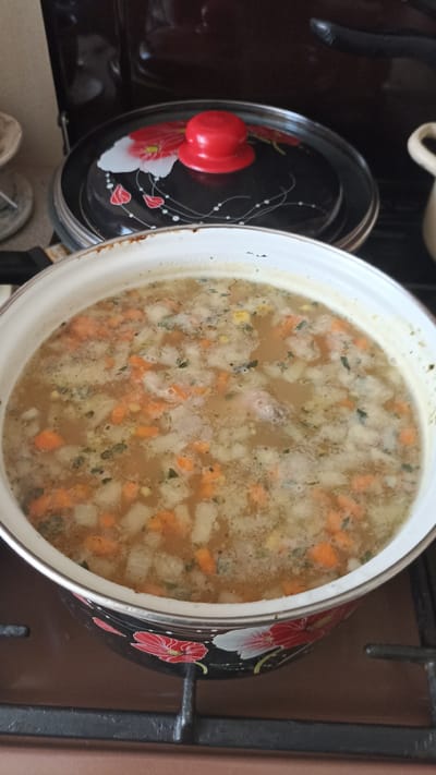 A close-up overhead shot shows a pot of soup simmering on a gas stove. The soup is a light brown color and contains visible chunks of carrots, potatoes, and herbs. The pot itself is white with a decorative floral pattern along the bottom edge, featuring red and white flowers on a black background. A black pot lid with a red knob is partially visible behind the soup pot. The stove is black with silver grates. The background shows a dark brown countertop and a portion of a dark appliance. There are no people or animals in the image. The scene is likely inside a residential kitchen.