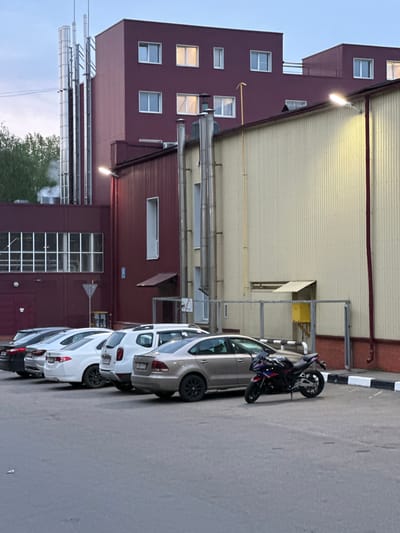 The image shows a parking area adjacent to a large industrial building. The building is characterized by a combination of maroon and beige siding, with visible windows and exterior piping. Several cars are parked in a row, ranging in color from white to silver, and a motorcycle is parked near the building. The location is likely in Reutov, Russia, based on the prompt. The environment appears to be urban-industrial. There is no clear indication of human presence or activity visible in the image.