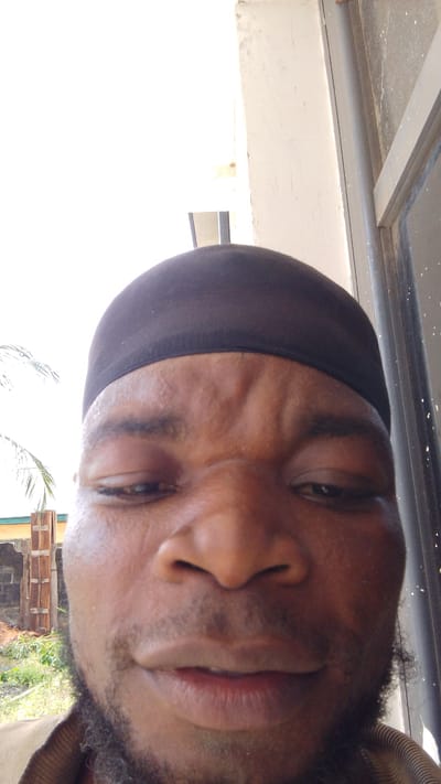 The image is a close-up selfie of a man with dark skin, a short beard, and a black head covering. He appears to be frowning slightly. The background shows part of a building with white walls and a window frame. Outside, there is greenery and a fence. The location is stated to be Enugwu-Ukwu, Nigeria.