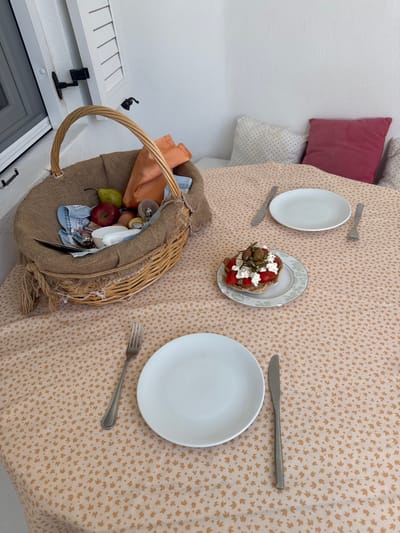 A high-angle, medium shot captures a table set for a meal, likely in Naousa, Greece, given the setting. The table is covered with a peach-colored tablecloth featuring a subtle, repetitive pattern of small, light brown leaf-like shapes.

Dominating the left side of the frame is a rustic woven basket, likely made of straw or wicker. It is filled with various items, including a bright red apple, a light green pear, eggs, and what appear to be small packets or wrappers. An orange fabric napkin is also visible within the basket. The basket has a sturdy, curved handle made of woven material and is lined with a burlap-like fabric, with fringed edges.

To the right of the basket, a small, round appetizer or snack is presented on a plate. It appears to be a piece of bread topped with crumbled white cheese, diced red tomatoes, and dark green olives.

Two place settings are arranged on the table. The setting closest to the viewer includes a white dinner plate, a silver knife to its right, and a silver fork to the left. Another white dinner plate is positioned further back on the table, with a silver fork to its right and a silver knife to its left.

In the background, a white wall is visible, with two pillows placed against it. One pillow is a muted pink color, and the other is white with a subtle dotted pattern. The top left corner of the image shows a portion of a white window frame with shutters. The overall impression is one of a simple, outdoor dining setting, possibly on a balcony or patio.