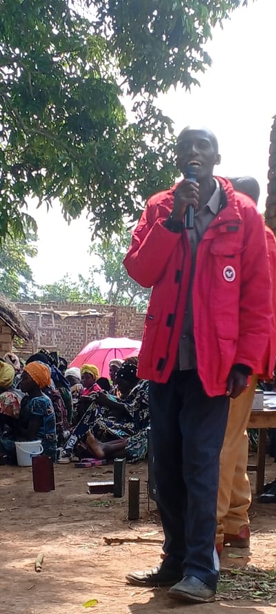The scene depicts an outdoor gathering in Fizi, Democratic Republic of the Congo, under bright daylight. Centrally, a dark-skinned adult male stands, holding a black microphone to his mouth as if speaking. He wears a red collared jacket with a white bird emblem on the left chest, a light-colored collared shirt, dark blue trousers, and dark closed-toe shoes. To his immediate left, a group of approximately 15-20 individuals, predominantly women and some children, are seated on the bare, light-brown dirt ground. They are dressed in various colorful patterned fabrics and headwraps, with one individual wearing an orange knitted cap. A red open umbrella is visible among the seated group, providing shade. On the ground near them are a small white plastic bucket and several dark rectangular objects, resembling books. In the background, structures made of rough brickwork and wooden elements are visible, partially obscured by a dense green tree canopy. A bright sky is seen above the foliage. A second individual, wearing light brown trousers, stands partially visible to the speaker's right. The ground is mainly bare earth, scattered with minor debris and a single green leaf.