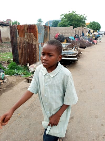 A young boy, appearing to be of African descent, walks on a dusty, unpaved road in Pantame, Nigeria. He is wearing a light green, short-sleeved collared shirt with a checkered pattern and dark trousers. The boy is looking to his right and his left arm is extended forward as he walks. Behind him, a makeshift fence of corrugated metal sheets separates the road from a cluttered area with grass, debris, and plastic bags. Further in the background, more people are visible near piles of scrap metal and various items, with a brick wall construction partially visible and trees and buildings in the distance. The overall scene suggests a busy, possibly informal market or residential area.