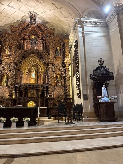 The image depicts the interior of the San Isidro Church in Lima, Peru. The focal point is the ornate, gilded altar with a central statue of the Virgin Mary holding the infant Jesus. Flanking the central statue are smaller statues of saints. The altar is intricately carved wood with gold embellishments. In the foreground, a marble altar rail is visible with several white floral arrangements. To the right of the altar, a separate, darker wooden pulpit is situated, featuring a statue of Mary on top, adorned with flowers. The church interior is illuminated by overhead lighting, casting soft shadows on the textured ceiling and stone walls. The scene is devoid of people, suggesting the image was captured during a quiet period.
