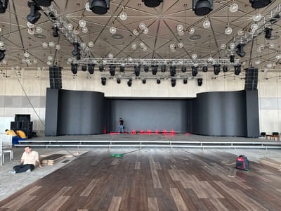 This image shows an indoor event venue, likely under setup or renovation, in Baku City, Azerbaijan. A large stage area dominates the background, featuring a dark, curved backdrop and a raised platform with rows of red stage lights. Above the stage, a complex rigging system suspends numerous black spotlights and decorative glass light fixtures from a geometric ceiling. In the foreground, a man in a light-colored t-shirt and dark pants is sitting on the floor, looking towards the camera. He appears to be taking a break. To his left, a stack of dark wooden planks, likely flooring material, lies on the ground. Further left, two individuals in yellow shirts and blue pants are standing near a stack of large black speaker cabinets. The floor is a mix of rough, unfinished concrete and newly laid dark wooden planks. The overall scene suggests an active preparation phase for an event.