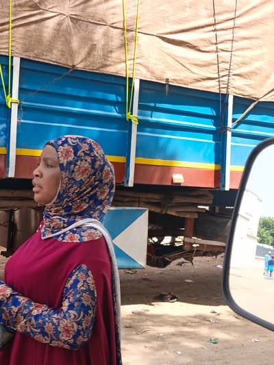 The image shows a woman wearing a hijab and a maroon dress with floral sleeves, standing in front of a large blue truck with a canvas cover. The truck has yellow and red stripes along its side. Yellow ropes are attached to the truck, extending upwards and outwards. The woman is looking to the left with a neutral expression. She is in profile.
In the background, there is a white building on the right, and a reflection in a rearview mirror shows two men in blue clothing walking in the distance. The ground is dusty and appears to be a road or an open area. Some trash is scattered on the ground.
The scene appears to be outdoors, possibly in a market or a roadside area in Fagge A, Nigeria. The lighting suggests it is daytime, with bright sunlight. The overall atmosphere is that of a typical day in a busy area. No visible text is present in the image. The focus is on the woman and the truck, with the surrounding environment providing context.