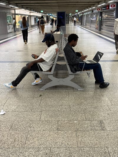 The image depicts a subway station platform in Duisburg, Germany. Two men are seated back-to-back on a metal bench. The man on the left, wearing a white t-shirt, black jeans, and a baseball cap, is looking at his phone. The man on the right, wearing a patterned jacket, dark jeans, and black shoes, is looking at a laptop and holding a phone. In the background, other passengers are walking along the platform, and shops are visible on the right side of the station. The platform floor is tiled with a granite-like pattern.