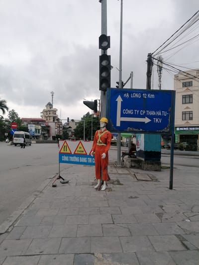 The image depicts an urban street corner or intersection in Ha Tu Ward, Vietnam, under an overcast sky during daytime. In the immediate foreground, on a paved sidewalk, a mannequin dressed as a construction worker stands. It wears an orange jumpsuit with reflective stripes and a yellow hard hat.
Beside the mannequin, a temporary A-frame sign with yellow and red triangles warns of ongoing work. To the mannequin's right, a prominent blue directional sign points towards "HẠ LONG 12 Km" (Ha Long 12 Km) with an upward arrow and "CÔNG TY CP THAN HÀ TU-TKV" (Ha Tu Coal Joint Stock Company - TKV) with a rightward arrow. Traffic lights are mounted on poles next to this sign.
In the background, a bustling street features various buildings, including a distinctive multi-story structure with a domed roof and a telecommunications tower on top. Trees and other urban foliage are also visible. A white minivan and a pedestrian are seen on the road. Numerous overhead utility cables are tangled across the scene. The sidewalk consists of grey square paving slabs, some of which appear worn.
Visible text includes:
* On the construction sign: "ĐI CHẬM" (Slow Down) and "CÔNG TRƯỜNG ĐANG THI CÔNG" (Construction Site in Progress).
* On the blue directional sign: "HẠ LONG 12 Km" and "CÔNG TY CP THAN HÀ TU-TKV".
* On building signs: "DISMY", and partially visible "CHĂM SÓC SỨC KHỎE" (Health Care) and "NHÀ THUỐC THANH BÌNH" (Thanh Binh Pharmacy).
