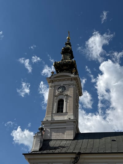 This image displays the upper section of a prominent church tower against a vibrant blue sky with scattered white cumulus clouds. The structure is the Name of Mary Church (Katedrala Imena Marijinog), a Roman Catholic church located on Trg slobode (Freedom Square) in Novi Sad, Serbia.
The central subject is the light-colored stone or stucco tower, which features a circular clock with a dark face and white Roman numerals, indicating a time approximately between 10:10 and 10:15. Below the clock is a recessed, rectangular window fitted with dark louvers or shutters. Ornate architectural details, including cornices and moldings, adorn the tower.
Crowning the tower is an elaborate, multi-tiered spire. The lower part of the spire is dark with visible gilded ornamentation, and the year "1903" is inscribed on one of its dark sections. The spire tapers upwards, culminating in a golden orb topped by a small, golden cross.
To the right of the tower, a portion of the church's dark, possibly metal, roof is visible, sloping downwards. Strong, direct sunlight illuminates the scene, casting subtle shadows on the left side of the tower and highlighting the architectural details. No people or other active subjects are present in the frame.