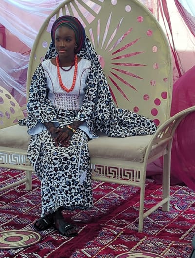 This image, captured in Unguwar Rimi, Nigeria, features a woman seated in a decorated setting, likely for a cultural event or ceremony. The central subject is an African woman dressed in traditional attire. She wears a white top with a patterned front, covered by a flowing garment adorned with black, white, blue, and reddish-brown floral or leaf patterns. Her head is wrapped in a colorful textile showcasing blue, green, and orange stripes. A prominent orange coral bead necklace graces her neck. Her hands, resting in her lap, display dark, intricate designs, possibly henna, and a multi-colored bracelet on her right wrist. She wears black sandals.
She is seated on a light-colored, possibly off-white or beige, ornate chair with a distinctive fan-like cut-out design on its backrest and geometric patterns on its armrest. The background is adorned with draped fabrics, including sheer white and solid pink/magenta materials, creating a festive atmosphere. The floor is covered by a richly patterned red rug with intricate white and blue geometric designs. A circular, woven mat or tray, also patterned, is visible on the rug to the right. The bright lighting suggests it is daytime. The woman maintains a calm and composed expression, appearing to pose for the photograph. No visible text is present in the image.