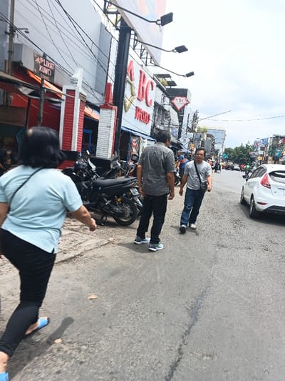 An outdoor street scene shows three people walking on a paved road. A woman in a light blue top and black pants is on the left, her back facing the viewer. Two men are walking to the right of her, one in a gray shirt and dark pants, and the other in a light gray shirt and blue jeans. Parked motorcycles and a white car are visible on the roadside. Buildings with storefronts line the street. The sky is overcast.