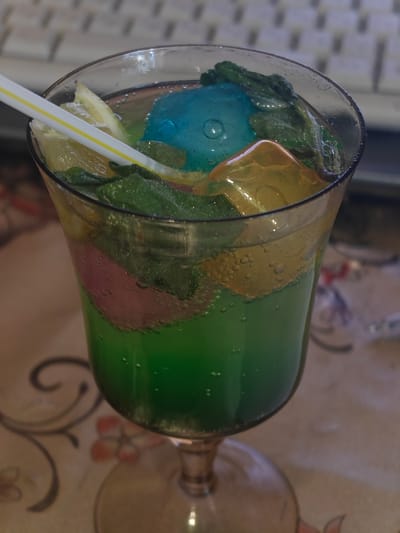 The image shows a close-up, high-angle view of a colorful drink in a glass. The drink is a vibrant green with layers of yellow and pink, and it contains various additions. Visible elements include:
* **Ice:** There are several pieces of ice, including a large, translucent blue ice sphere or cube, and several amber-colored, irregularly shaped ice cubes.
* **Fruit/Garnish:** Slices of what appear to be lemon or lime are present, along with green leaves, likely mint, scattered throughout the drink.
* **Liquid:** The drink itself is a bright green, with visible bubbles suggesting it is carbonated. There are also hints of other colors within the liquid, possibly from syrups or other ingredients.
* **Straw:** A white and yellow striped straw is inserted into the drink, resting against a lemon slice.
* **Glass:** The drink is served in a clear glass with a stemmed base. The outside of the glass shows condensation.
* **Background:** The background is out of focus but appears to be a surface with a patterned tablecloth featuring floral or swirling motifs. Behind the drink, a portion of a keyboard is visible.
The lighting suggests it might be indoors, possibly in daylight or under artificial light. There are no people visible in the image. The overall impression is of a refreshing, visually appealing beverage. The context provided (Hadiach, Ukraine) indicates the geographical location of the image, but no specific visual cues within the image confirm this. There is no visible text in the image.