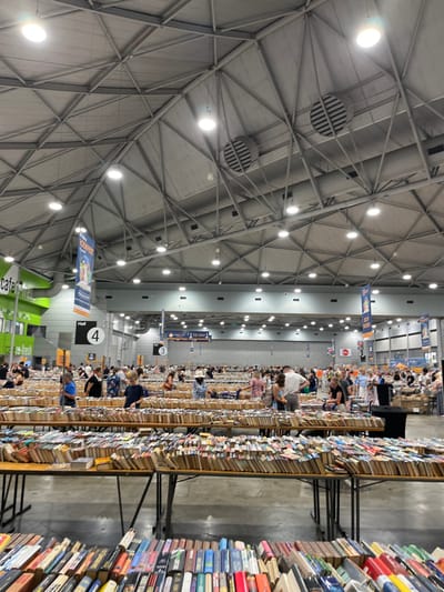 This image captures a large indoor book sale or book fair, likely set within an exhibition hall in South Brisbane, Australia. The scene is bustling with numerous people, predominantly adults, browsing an immense quantity of books.

The venue features a high, industrial-style ceiling constructed from exposed metal trusses, with numerous circular light fixtures providing bright, even illumination. Large ventilation grates are also visible on the ceiling. Below, long rows of tables are laden with books, stacked both upright and in piles, creating extensive aisles for visitors to navigate. Books are displayed on sturdy metal tables, with some overflowing onto the floor in organized stacks.

People are seen scattered throughout the hall, with many bent over or standing close to the tables, carefully examining books. The atmosphere appears busy but focused, with attendees engrossed in searching for titles.

Notable visible text includes a green "cafe" sign in the foreground, and further back, signs indicating "TO PAY," "3," and "Hall 4," suggesting an organized layout for the event. A banner partially visible in the distance also contains the word "experience," possibly referring to a specific event or brand. The sheer volume of books and the number of attendees indicate a popular and large-scale event. The indoor setting, artificial lighting, and absence of natural light suggest it is either daytime or evening, but well-lit indoors.