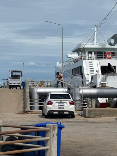 A white sedan is parked on a ramp leading to a large white ferry docked at a pier in Don Sak, Thailand. The sedan has its rear facing the camera. A man in an orange shirt and dark shorts is standing on a metal walkway on the side of the ferry, holding a rope. A small white truck is parked further up the ramp. The sky is overcast with clouds. There are large pipes and a red life preserver visible on the ferry. Blue and black trash bins are located in the foreground. The scene appears to be a ferry terminal or loading area.