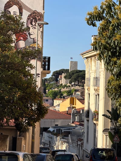 The image, rotated to its correct upright orientation, captures a sunlit street scene in what appears to be a historic European town, specifically Cannes, France, as indicated by the context. The perspective looks up a gently ascending street, flanked by buildings and trees, culminating in a prominent stone tower on a hilltop.

In the immediate foreground on the left, a vibrant, colorful mural adorns the side of a building. It depicts two stylized human faces in close proximity; one, possibly male, has brown, curly hair and a subtle horn-like detail, while the other, possibly female, features long, flowing hair. A lush tree with dark green leaves and small yellow, spherical fruits (resembling loquats) partially obscures the lower part of this mural. On the right side of the foreground, several cars are parked along the street. A white triangular road sign with a red border and a black curved arrow, warning of "DANGER," is visible above the vehicles.

The midground features a dense array of buildings climbing the hillside. On the right, a tall, light-cream colored building stands out with ornate architectural details, including decorative balconies and numerous closed shutters. Further up, a patchwork of red-tiled roofs, yellow, white, and grey facades, and scattered green trees creates a textured urban landscape.

At the top of the hill, in the background, a distinctive stone tower or castle-like structure commands the skyline. It is surrounded by dense, dark green foliage. A small flag, displaying horizontal stripes of what appear to be red, white, and blue, flies atop this distant tower.

The clear, bright blue sky and pronounced shadows indicate that the photograph was taken on a sunny day, likely during the mid-morning or afternoon. The scene is calm, with no people actively visible. The combination of historic architecture, vibrant mural, and Mediterranean vegetation evokes a picturesque and characteristic European town setting.