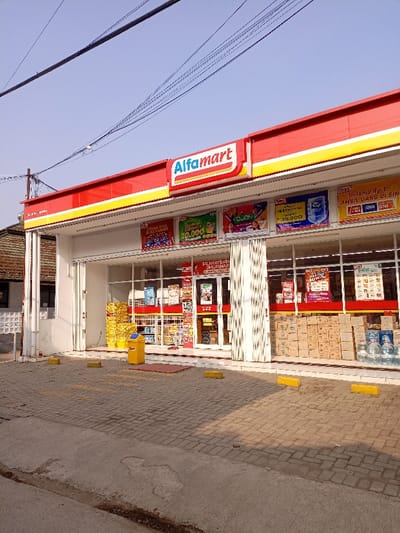The image shows an Alfamart convenience store in Katapang, Indonesia. The store is located at the coordinates -6.984, 107.554. The store is a one-story brick building with a red and yellow awning. The storefront is made of glass, and there are several signs hanging from the awning. There are several rows of packaged goods and drinks on shelves inside the store, some of which are visible through the windows. There is a yellow trash can in front of the store, on the paved parking lot, and there are three yellow bollards to the left and right of the entrance. The street in front of the store is paved and there is a small, gray strip of sidewalk in front of the store. In the distance, a dark-colored building is visible. The sky is blue and there are power lines overhead. There are no people visible in the image.