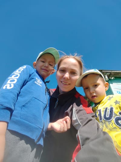 A low-angle, ground-level outdoor photograph shows a woman and two young boys taken against a clear blue sky. The woman, with her hair pulled back and a visible smile, is in the center, her face partially obscured by the children. She is wearing a dark jacket with red and grey accents. To her left, a boy in a blue hooded jacket with white lettering and an Adidas logo, and a light blue cap, looks towards the camera. To her right, a boy in a yellow hooded sweatshirt with a pattern and a white and blue cap, is looking directly at the camera. Behind them, a portion of a light blue wooden building is visible. The scene suggests a casual, sunny day outing, possibly in a residential area, given the presence of the building. The image is likely taken in Chukhlem, Russia, based on the provided location context.
