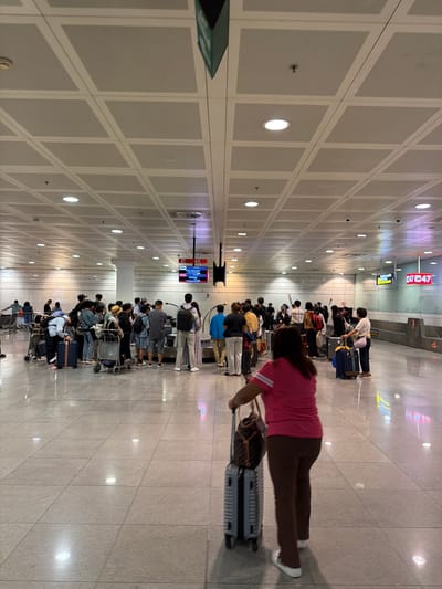 The image shows a crowded airport baggage claim area. In the foreground, a woman in a pink shirt and brown pants is walking with a rolling suitcase. The floor is tiled and reflects the overhead lights, which are housed in a grid-like ceiling structure. In the midground, a long line of people, many with luggage, are waiting. Several baggage carousels are visible in the background, along with a digital display board showing flight information. Signs for "GATE 47" are also visible. The overall environment suggests a busy travel hub, likely within El Prat de Llobregat, Spain, given the context of the provided crops hinting at airport identifiers.