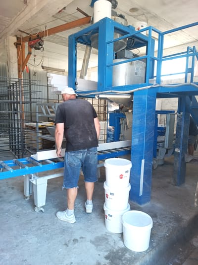 In an industrial setting, a man wearing a black t-shirt, denim shorts, a white cap, and white sneakers stands facing away from the camera, looking at a white, long, rectangular object on a conveyor belt. The object appears to be a piece of trim or molding. The conveyor belt is blue and grey and moves horizontally. Behind the man, a large, blue industrial machine with various metal components and chutes is visible. To the right of the machine, a stack of white plastic buckets sits on the concrete floor. The environment is a large, open space with a concrete floor, exposed beams and pipes on the ceiling, and metal shelving units filled with items in the background. The overall impression is a manufacturing or processing facility.