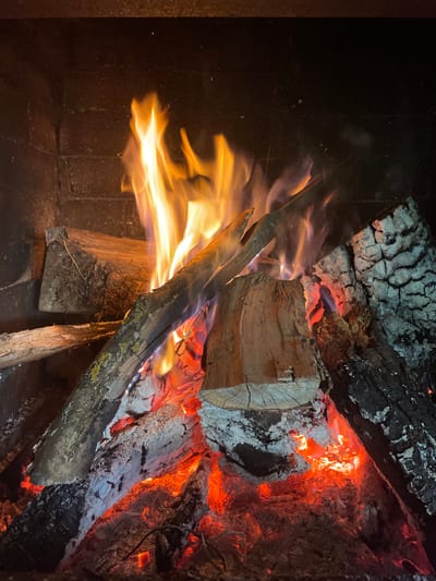 The image depicts a fire burning inside a fireplace. Several logs are arranged to fuel the fire, with active flames visible. The logs range from newly added pieces to heavily charred remains of previous fires. The fireplace interior appears dark and soot-covered. The location in Gryaznoye, Russia, is not discernible from the image.