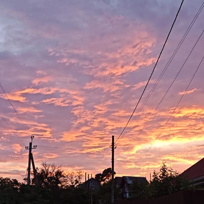 The image depicts a sunset over urban settlement Shchelkino, Ukraine. The sky is filled with a dramatic display of clouds, colored in hues of pink, orange, and purple, indicative of a sunset. Silhouetted against the vibrant sky are power lines and utility poles, crossing diagonally through the frame. Below the sky, the tops of trees and houses are visible, also appearing as dark silhouettes against the bright sky. The landscape suggests a residential area with some vegetation.