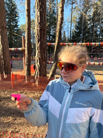 A young person wearing a light blue and white Glissade jacket and orange-tinted sunglasses stands outdoors, holding a small, pink object in their right hand. The background includes trees, orange safety netting, and structures. The location is Lesnoye, Russia.