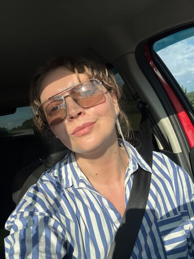 A close-up, slightly low-angle, selfie taken from inside a vehicle shows a woman with light brown hair pulled back, wearing large, aviator-style sunglasses with a pinkish tint. She is looking directly at the camera with a slight smile. She has on a blue and white striped button-down shirt. A silver, dangly earring is visible on her left ear. Sunlight streams in from the right, casting a shadow across her face and neck. The interior of the car is visible, including a red door frame, a black seatbelt across her chest, and the ceiling of the car. Through the windshield and side windows, a blurred, outdoor scene is visible, suggesting the vehicle is in motion. The image quality is consistent with a smartphone photograph. The location is specified as Polyakovo, Russia, which is an urban or suburban area, but no specific landmarks within the city are discernible from the image itself.