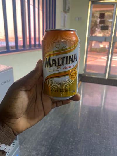 A dark-skinned right hand, adorned with a light-colored beaded bracelet on the wrist, holds an orange, white, and gold 33cl Maltina Classic non-alcoholic malt drink can. The can's label indicates it is "MULTI VITAMIN ENRICHED" and "FORTIFIED WITH CALCIUM AND VITAMINS." The scene is indoors, located in Kaduna South, Nigeria. In the background to the left, a window with vertical blue blinds allows warm, pinkish-orange light to enter. Partially visible to the far left is a white appliance labeled "CWAY." To the right, a glass door reflects ambient light and shows indistinct outdoor elements beyond. The flooring appears to be a light-colored, reflective surface.
