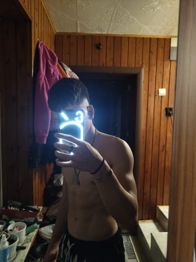 The image displays a mirror selfie of an individual, likely male, captured from the chest up. The person is shirtless, revealing a muscular build, and wears dark shorts. They have short, dark hair. Their left hand holds a smartphone, whose screen emits a bright blue glow, displaying a stylized unhappy or sad emoji face. The individual wears a black bracelet on their right wrist, a dark ring on a finger of their left hand, and a necklace with a pendant.

The indoor setting is characterized by a combination of wooden wall paneling and textured white wallpaper. Below the wooden wall, a dark wooden surface holds a folded pink blanket or towel. A dark doorway is visible in the wooden wall, along with a white light switch and a black electrical outlet. In the bottom right of the reflection, various household items, including white cups and a pump bottle, are present on a surface.

The scene is dimly lit, suggesting the photograph was taken in the evening or at night, with the phone's bright screen serving as a primary light source. The individual's face is not visible. The location context is Yambol, Bulgaria.