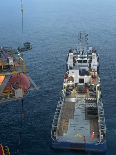 This image captures an aerial view of two vessels at sea. The main subject is a large vessel, likely a supply ship or offshore support vessel, with a distinctive blue hull and a white superstructure. The deck is filled with various equipment, including storage containers, ropes, and machinery. Two individuals in orange safety suits are visible on the deck, suggesting work is in progress or has recently occurred. The vessel has the name "VEYOLA BLUE" visible on its superstructure.
In the foreground, another vessel, possibly a smaller platform or lifeboat davit system, is partially visible. It features an orange lifeboat and a green crane structure. A cable stretches between the two vessels, suggesting a transfer or connection is being made. The sea is calm with small ripples, indicating a tranquil environment. The lighting suggests it is either dawn or dusk, with a soft, diffused light. There are no specific indicators of weather conditions, but the calm sea implies fair weather. The location is unknown but has been contextually linked to Italy. No overt emotions are conveyed, but the presence of personnel in safety gear points to an operational setting.