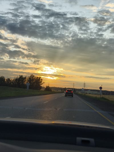 This image depicts a sunset scene on a highway, likely in or near the city of Kuraskovo, Russia. The sky is filled with dramatic clouds, with rays of sunlight breaking through, illuminating the scene with a warm, golden glow. A dark-colored sedan is traveling on the right lane of the two-lane highway. The road ahead curves slightly to the left. On the right side of the road, there are fields and power lines. On the left side, there are trees and vegetation. The foreground shows the dashboard of a vehicle, indicating the photo was taken from inside a car. The time of day is dusk, given the sunset lighting.