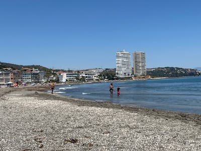 The image depicts a beach scene in San Roque, Spain, under a clear blue sky. In the foreground, a pebble beach stretches towards the calm, blue sea. Several people are visible on the beach and in the water. A woman and a child are standing in the shallow water, facing each other. Further down the beach, a lone person stands near the water's edge. In the middle ground, a line of buildings, including apartment complexes and hotels, forms the urban backdrop to the beach. Two tall, white apartment buildings with balconies are prominent features in the cityscape. The coastline extends into the distance, with hills covered in greenery and more scattered buildings. The overall impression is of a sunny, populated beach destination.
