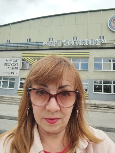 A woman with auburn hair and large, patterned glasses is posing for a selfie in front of a building. The building has a light-colored, corrugated metal facade with a row of windows. Below the windows, there is a sign with yellow and black stripes, and a white banner with text in a foreign language. Above the windows, there is a large blue sign with white letters. The woman is wearing a light-colored jacket over a red top. The environment appears to be outdoors, with a cloudy sky visible.