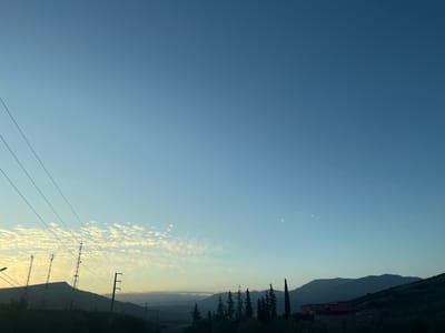 The photograph captures a landscape near Ouaouizert, Morocco, at dawn or dusk. The sky transitions from a light blue near the horizon to a deeper blue overhead. Altocumulus clouds are scattered in the distance.

Mountains form the horizon, their silhouettes varying in height and shape. In the foreground, there are evergreen trees and a building. Power lines stretch across the left side of the frame. Several communication towers are visible on a hill.