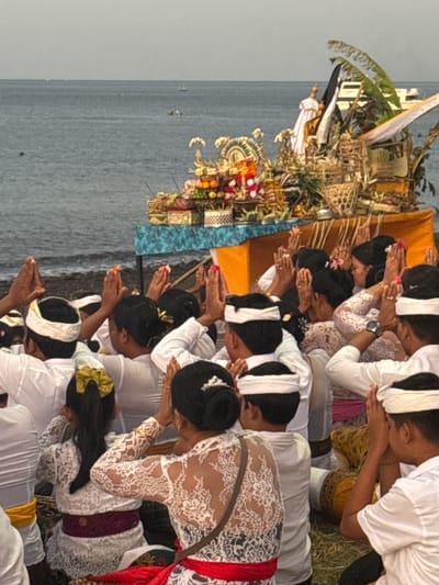 The image shows a group of people in traditional white clothing with headbands praying with their hands clasped together. They are facing a decorated altar with offerings, including fruits and flowers, set up on a table by the sea. The altar is adorned with a white statue, flags, and woven baskets. The scene appears to be a religious ceremony or ritual taking place outdoors, near the ocean. The background shows the calm sea and the sky, suggesting an early morning or late afternoon setting. The location is identified as Abang, Indonesia.