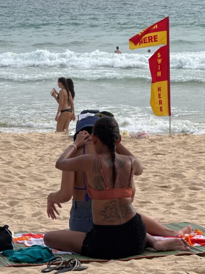 This image depicts a beach scene with several individuals. Two women are prominently featured in the foreground, with one lying on a colorful striped towel on the sand, and another woman in the background, partially submerged in the ocean. The woman on the towel is wearing a coral-colored bikini top, black lace shorts, and has extensive tattoos on her back and arms. The woman in the ocean is wearing a dark bikini. Further in the background, another person is visible in the water.
The setting is a beach with sandy ground and rough, choppy waves rolling in from the ocean. Two red and yellow flags are planted in the sand near the water's edge, indicating a swimming area. The flags have text visible on them. One flag has "HERE" printed in yellow on a red background, and another flag has "SWIM" in red lettering on a yellow background. The image appears to be taken during daylight, with bright lighting, suggesting a warm, possibly tropical environment. The overall mood is relaxed and leisurely, characteristic of a beach day. The location is identified as Rawai, Thailand, which aligns with the tropical beach setting.