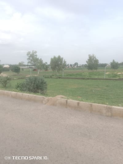 The image depicts an outdoor scene with a paved road in the foreground, bordered by a concrete curb and a grassy area. Beyond the grass, there are several trees, some scattered and some forming a line. In the background, there are buildings and more trees, suggesting a residential or semi-urban environment. The sky is overcast and appears bright, indicating a cloudy day. The overall impression is one of a tranquil, undeveloped area, possibly on the outskirts of a town or city. The "TECNO SPARK 10c" watermark in the bottom left corner indicates the device used to capture the image. Based on the description provided, the scene is located in Kason, Nigeria.