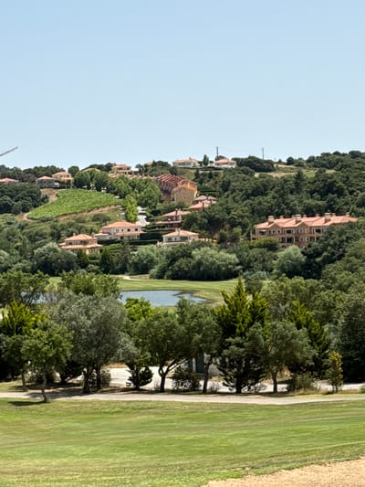 This image depicts a panoramic view of a residential area nestled within a hilly, green landscape, likely located in the vicinity of Torres Vedras, Portugal. The scene is dominated by lush vegetation, including numerous trees and vineyards, suggesting a rural or semi-rural setting. Several multi-story residential buildings with red-tiled roofs are scattered across the hillsides, appearing to be modern villas or apartment complexes. A body of water, possibly a small lake or pond, is visible in the mid-ground, surrounded by trees. In the foreground, a well-maintained golf course or park area with vibrant green grass is present, with a paved pathway winding through it. The sky is clear and blue, indicating fair weather. There are no people or animals visible in the frame. The overall impression is one of a serene and affluent residential development integrated into a natural environment.