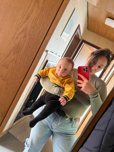 A mirror selfie taken in an indoor residential setting within the city of Wüstenrot, Germany, captures an adult female holding an infant. The female, positioned on the right side of the frame, has dark hair pulled back and wears a long-sleeved olive-green top, light-wash blue ripped jeans, and a dark belt. She holds a red-cased smartphone in her right hand, directed towards the mirror to capture the image. Her left arm supports an infant, who is dressed in a yellow long-sleeved top with a partially visible "YOU" inscription and black pants with matching socks. The infant looks directly forward with wide eyes. The reflected background includes a light brown wooden door or panel on the left, textured off-white wallpaper, a framed rectangular object mounted on the wall, and a white rectangular vent. The ceiling features light brown wooden paneling and a white rectangular light fixture. A section of a dark blue coat is reflected on the bottom right of the image.