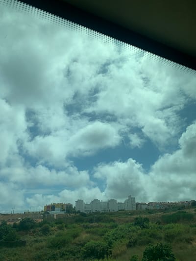 This is a vertical, landscape photograph taken from inside a vehicle, looking out through the windshield. The image depicts a cloudy sky with patches of blue visible through the cumulus clouds. To the right of the sky, there is a hillside covered in green vegetation and trees. Along the crest of the hill, a line of multi-story buildings is visible, some appearing to be residential complexes. The architecture suggests a Mediterranean or Southern European style. The lighting indicates it is daytime, likely late morning or afternoon, given the bright sky and shadows. The overall mood is serene, capturing a view of urban development against a natural backdrop. No people are visible in the frame. The location is identified as Lisbon, Portugal, which aligns with the visual cues of the architecture and landscape. The time of day and weather are both contributing to a pleasant, though overcast, atmosphere.