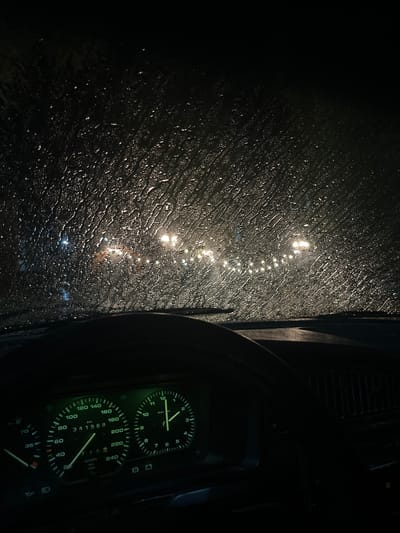 The image is a dimly lit, vertical shot taken from inside a vehicle at night. The foreground shows a dashboard with illuminated gauges, including a speedometer and a clock. The windshield is covered in streaks of rainwater, distorting the view of the outside. Beyond the windshield, through the rain, a line of blurred lights is visible, suggesting a street or an outdoor area with multiple light sources. The background is dark and indistinct. The scene is indoors, within a car, looking outwards on a rainy night. There are no people or distinct actions visible.