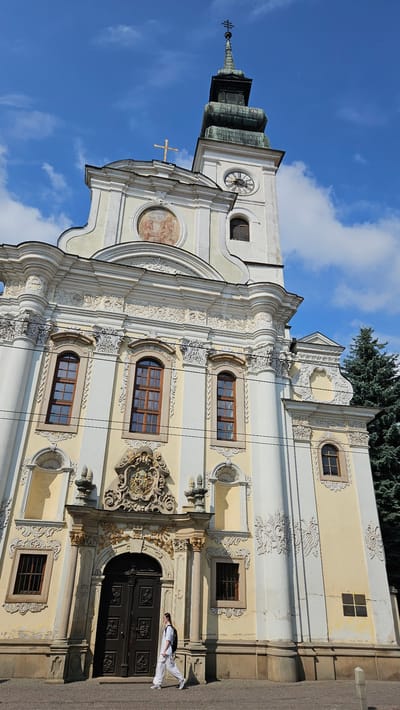 The image captures a low-angle, full-shot of the St. Nicholas Cathedral in Prešov, Slovakia. The cathedral is a prominent baroque-style building with a striking yellow facade adorned with intricate white stucco decorations. A tall, multi-tiered bell tower with a green, onion-shaped spire rises to the right of the main facade. The tower features a clock face. A golden cross is prominently displayed at the apex of the spire and another golden cross is situated above the central pediment.
In front of the cathedral entrance, a person, appearing to be male and dressed in white clothing and carrying a backpack, is walking from left to right. The person is the sole human subject in the frame. The entrance itself is a grand, dark wooden double door, framed by ornate golden sculptures and columns. Above the door, a complex coat of arms is displayed.
The sky is a vibrant blue with scattered white clouds, indicating a clear day. To the far right of the image, the dark green branches of a coniferous tree are visible, partially obscuring the side of the cathedral. Power lines are also visible in the mid-ground, crossing horizontally in front of the building. The ground in the foreground is paved with cobblestones. The overall scene depicts a sunny day with a focus on the architectural grandeur of the cathedral and the solitary figure passing by.