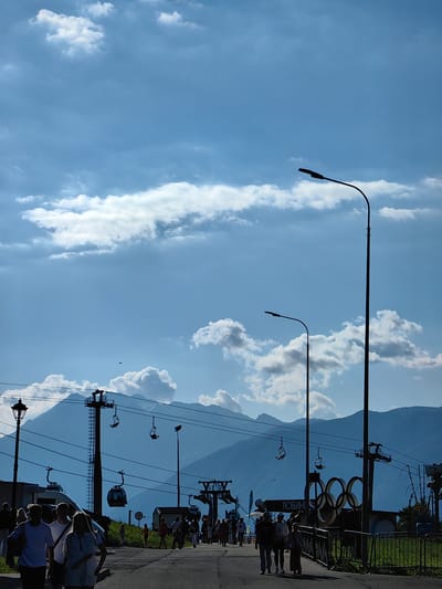The image depicts a scene at a mountain resort, likely during daytime. Silhouettes of people are walking along a paved path in the foreground, suggesting a leisurely stroll or an outing. In the background, a ski lift system with chairs and gondolas is visible, indicating the presence of mountains and a place for winter sports or scenic rides. The sky is a bright blue with scattered white clouds, suggesting fair weather.

Several notable objects are present. Tall lampposts with modern fixtures line the path. Prominently featured is a large display of the Olympic rings, a clear indication of the location's connection to the Olympic Games, likely the 2014 Sochi Winter Olympics given the geographical context. There is also a sign with Cyrillic text, reading "ПОБЕДЫ" which translates to "VICTORY". The overall mood appears to be peaceful and recreational, with people enjoying the outdoors and the mountainous scenery. The sun is casting shadows, implying it is either morning or afternoon. The scene captures a blend of natural beauty with man-made recreational facilities.