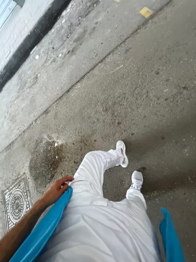 The image shows a section of a city street, likely in the Sarouja Municipality area of Syria, given the request. The ground is paved and shows signs of wear and discoloration. A person wearing white clothing and white sneakers is partially visible, from the legs up. The individual is holding a blue object, which appears to be a long, curved plastic or fabric item. A sewer grate is present in the lower right corner of the frame. The overall scene has a gritty, urban feel.