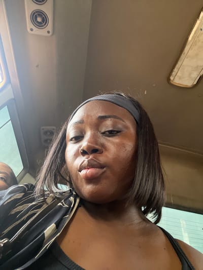 This is a selfie taken by a young Black woman inside a vehicle, likely a bus. She is wearing a grey headband and a black top. She has a nose ring. Her hair is dark and straight, falling around her face. The background shows the interior of the vehicle, including air vents and a window. The lighting suggests it is daytime, and the reflection in the window indicates an outdoor scene. The woman's expression is neutral, and she is looking directly at the camera. A second person's face is partially visible in the lower right corner of the frame. The scene appears to be within Kugbo, Nigeria.