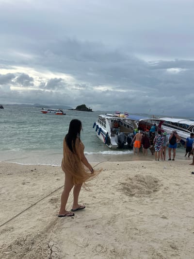 In Phru Nai, Thailand, a group of people are disembarking from a white and blue speed boat onto a sandy beach. The boat is positioned close to the shore, with its rear engines partially submerged in the shallow water. Several individuals, including a man in orange shorts and a woman in a colorful patterned dress, are seen stepping out of the boat onto the sand. Another boat, primarily white with orange accents and bearing the text "S.AREEL", is anchored further out in the choppy, green water. A woman with long dark hair, wearing a sheer tan cover-up over swimwear, stands on the beach facing away from the viewer, looking towards the boats and the sea. The sky above is overcast with heavy, gray clouds. In the distance, several islands with lush green vegetation are visible, shrouded in the hazy atmosphere. The beach itself is composed of light-colored sand with some scattered debris. A rope lies on the sand near the woman in the tan cover-up, extending towards the water's edge.